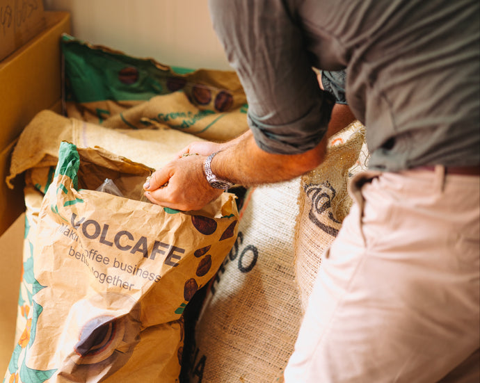 Person handling a Volcafe coffee bag in a storage area with other bags and boxes.